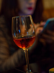 Rose wine glass with woman blurred in the background with cellphone. Woman waiting in a bar for a date while drinking. Brunette with phone in a restaurant alone.
