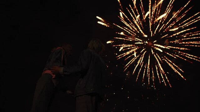 Young couple in love are standing outdoors against background of night sky and fireworks, back view. Couple of men and women stand against sky with fireworks