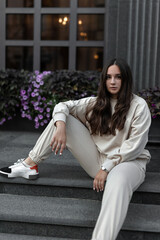 A girl in a white tracksuit on a city street on a sunny clear day walks posing for the camera sits on the steps of the hotel building near the column