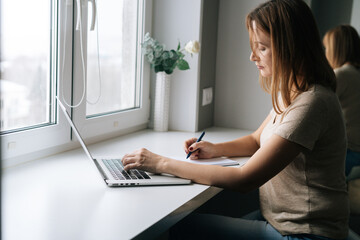 Side view of focused business woman writing making note in paper notebook sitting at windowsill with laptop, working from home office. Female freelancer using computer by window at workplace
