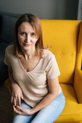 High-angle view of pretty blonde woman sitting in comfy yellow chair in dark room with modern interior and looking at camera, selective focus. Happy female resting in chair alone.