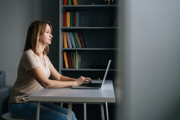 Attractive business woman working on laptop computer from home office sitting at desk in living room, looking on screen. Serious businesswoman studying online, web surfing information, shopping.