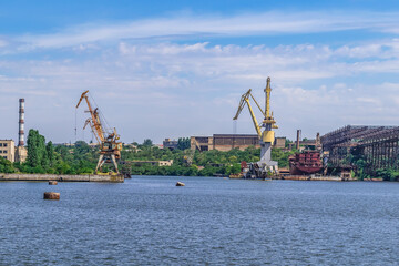 Industrial landscape of the Shipyard on the Inhul river in Mykolaiv, Ukraine. Port cranes and repair shops on the coastline with blue water in the foreground