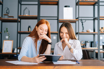 Portrait of two laughing young business women working using digital tablet at meeting desk with job documents at office. Business female colleagues using touchscreen computer for project discussion