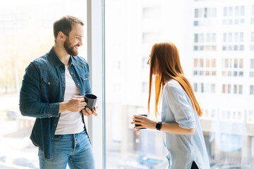 Two cheerful young man and woman colleagues with coffee cups standing by window in modern loft office and discussing business issues. Relaxed coworkers having conversation during coffee break.