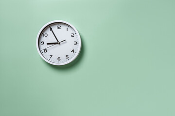 Wall clock hanging on the pale green wall with copy space. Round white clock with black hands. Five...