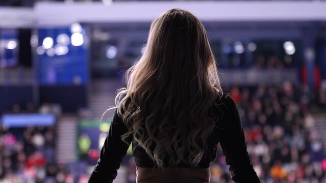 Blonde slim cheerleader with shiny pompoms dances to amuse audience and players at hockey game backside view slow motion