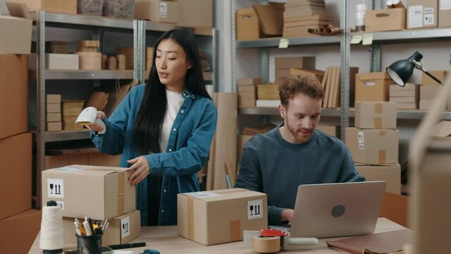 Cute asian woman talking with caucasian man while scanning barcode at the cupboard box and preparing delivery parcel box. Post service and small business concept.