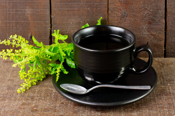 Tulsi or holy basil tea in black cup with fresh tulsi branch on wooden background. Ayurvedic medicine in India.
