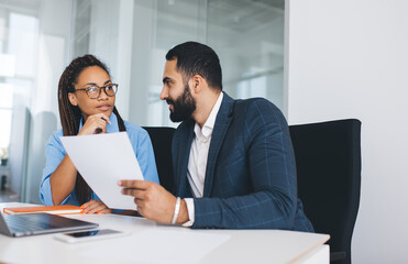 Young male and female colleagues discussing information during cooperation briefing in office interior, multicultural partners in formal clothes analyzing paperwork reports and documents in company