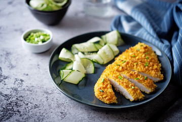Bread crumbs cheesy chicken breast with cucumbers in a plate