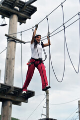 A young woman in a rope amusement park crosses a suspension bridge on a rope