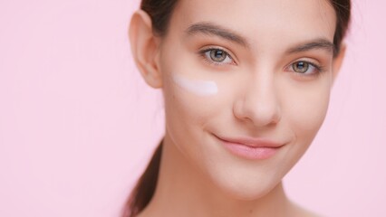 Obraz premium Close-up portrait of young attractive beauty model with perfect skin with cream smear on her face looks at the camera and smiles on light pink background | Skin care product advertising concept.