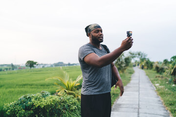 Dark skinned male tourist talking while shooting selfie influence vlog via Go Pro camera technology during summer trip at Bali with rice fields, young hipster guy using waterproof device in Indonesia