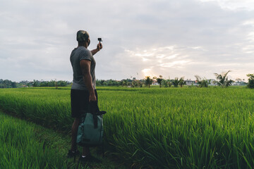 Back view of male influencer shooting vlog content via digital Go Pro technology - summer trip vacations in Indonesia, man with touristic backpack and waterproof camera making photo at rice fields