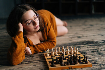 Attractive sexy young woman in elegant eyeglasses thinking about chess move lying on wooden floor...