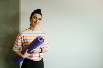Pretty young woman holding purple yoga mat near wall at home, copy space