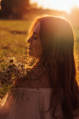 silhouette of a beautiful young girl with red curly hair in a dress standing with closed eyes and holding in his hands near the face of daisies at sunset side view close up defocus on the girl's face