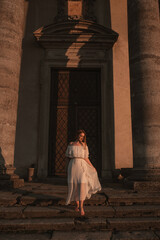 beautiful young girl with red curly hair in a long white dress barefoot walks up the old stairs near the old gray building with columns of ancient architecture at sunset, foreground