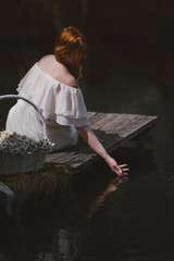 beautiful young girl with red hair in a white dress sits on a wooden pier and touches his hand to the water near her white straw basket with daisies, sunny day side view