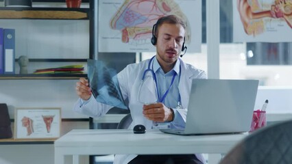 In modern room attractive man doctor in white medical coat with headphones use laptop consulting distance patient online in video chat. Hold x-ray. Slow motion