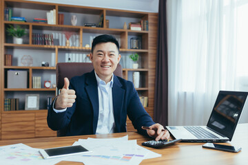 Portrait of Asian financier, businessman working in office, paperwork, looking at camera and smiling, showing thumbs up