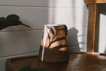 Fototapeta na wymiar Modern ceramic kettle in sunlight on rustic wooden chair and leaves shadow. Morning aesthetics