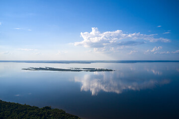 A full-flowing flat river with forested banks. There is a small island in the middle of the river. The white cloud is beautifully reflected in the water. Shooting from a drone.
