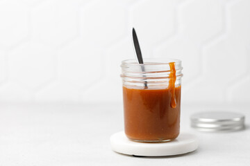 Salted caramel sauce in glass jar with lid on marble board, white background. Jar with Homemade caramel and delicious drop.