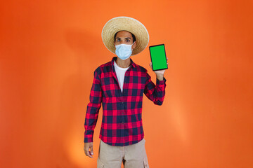 Black Man With Junina Party Outfit with Pandemic Mask Holding a tablet Isolated on Orange Background.