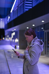 Young millennial woman using technology - holding and texting on mobile phone, or video chatting, or making video call using social media standing near office building at night with blue neon lights