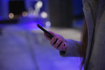 woman using technology - closeup hands holding and texting on mobile phone, or video chatting, or making video call using social media standing near office building at night with blue neon lights