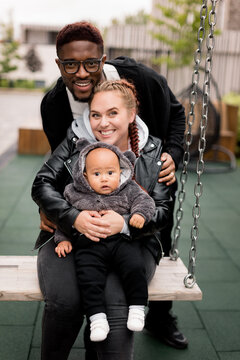 Portrait of stylish multiracial family with cute toddler at the park, smiling and hugging. Young beautiful parents with little son having a walk, looking at the camera, weekends outdoor concept