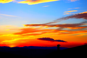 Fototapeta na wymiar Impressive sunset sky of a very bright red, yellow and blue color spectrum with peculiar clouds and silhouettes of a roof and a hilly valley in Monte Vidon Corrado
