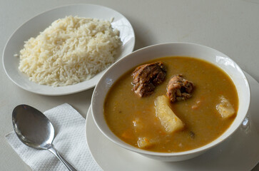 A white bowl of Dominican sancocho with yucca, chicken, pork, squash and more, served with white rice and a metal spoon.