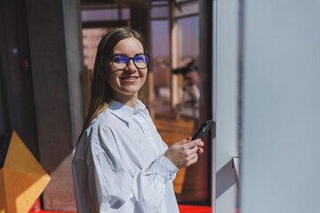 Smiling woman in glasses in casual wear watching a positive video on a phone connected to wi-fi, a female blogger in a white shirt against a large window, enjoying relaxation during remote work.