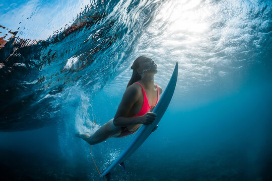 Surfer woman with surfboard dive underwater with under wave and sunlight in transparent ocean.