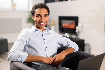 Smiling intelligent multiracial male freelancer in smart casual shirt using laptop for work online, sitting in the armchair and looks at camera in the coworking space
