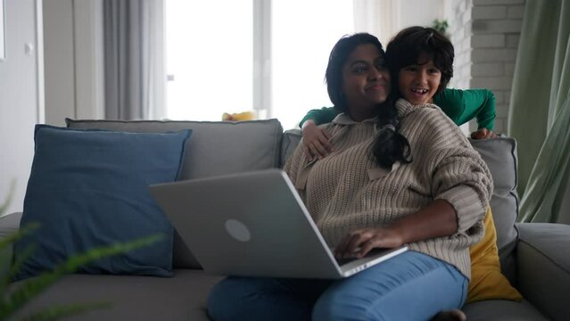 Indian mother using laptop and her little son is suprising her on sofa at home.