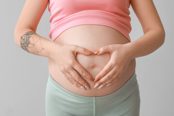 Pregnant woman making heart shape with her hands on belly against grey background, closeup