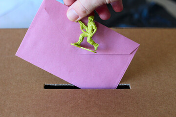 un soldat jaune et une enveloppe en papier rose tenu par une main, au dessus d'une fente d'urne vote