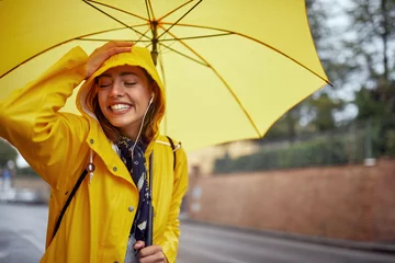 Fotobehang Close-up of a young cheerful woman with a yellow raincoat and umbrella who is in a good mood while walking the city on a rainy day and posing for a photo. Walk, rain, city © luckybusiness