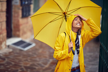 A young cheerful woman with a yellow raincoat and umbrella who is in a good mood while walking the city on a rainy day. Walk, rain, city
