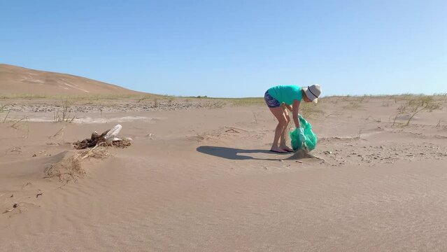 Mature woman in a white hat and green gloves collecting plastic garbage from de sand dunes in a green bag. 