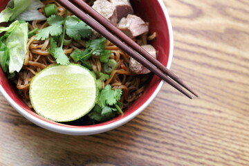 Stewed pork Noodle garnished with coriander, sliced spring onions and lemon wedges. Served on a walnut patterned wood table.
