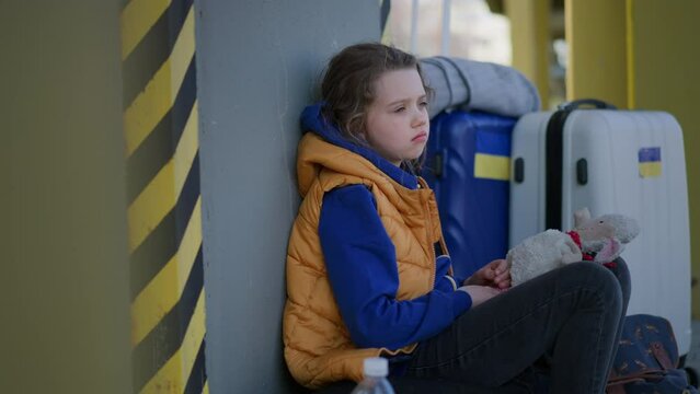 Sad Ukrainian immigrant child with luggage waiting at train station, Ukrainian war concept.