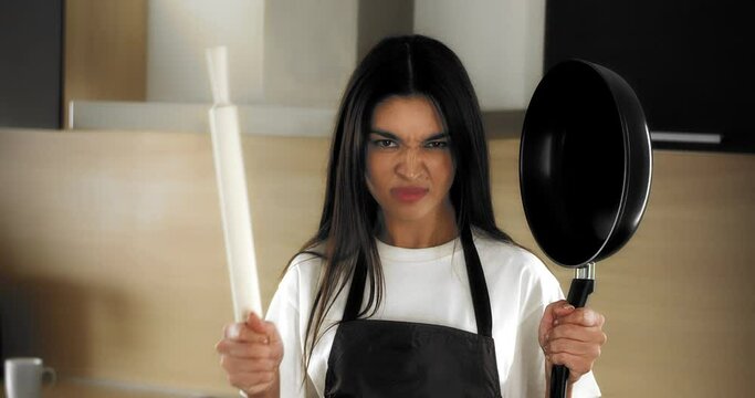 A disgruntled, tortured housewife in a white T-shirt and brown apron stands in the kitchen, threateningly brings a rolling pin and a frying pan to her face, tired of cooking