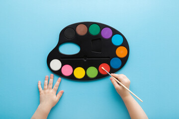 Baby hand holding paint brush and using colorful palette on light blue table background. Pastel...