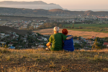 Fototapeta na wymiar An elderly couple sits on a mountain with their backs with a beautiful view of the mountains and the sea in the distance.