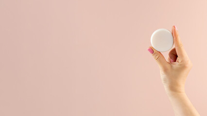 A woman's hand holds a round piece of soap on a light pink background. Banner
